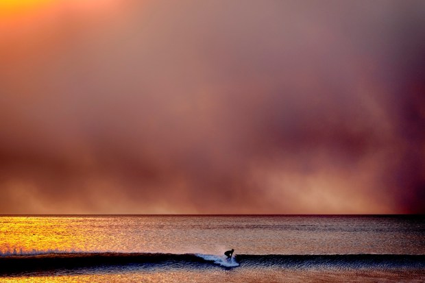 A surfer takes off on a wave in Santa Monica, Calif., during sunset under a blackened sky from the Palisades fire