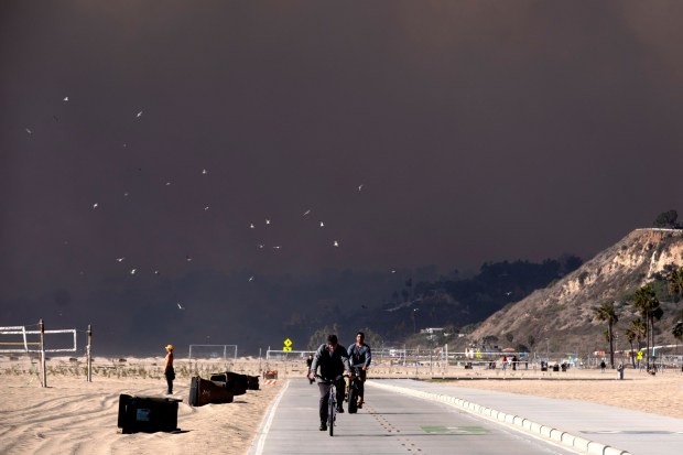 Bike riders make their way along the coast as large dark plume of smoke passes over the beach from a wildfire from Pacific Palisades