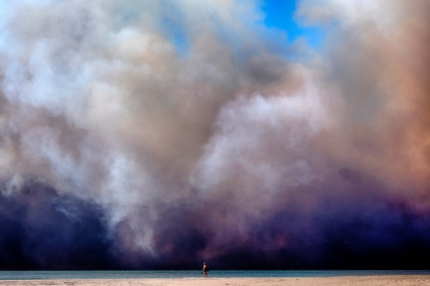 A lone beachgoer walks along the coast as a large dark plume of smoke passes over the beach from a wildfire from Pacific Palisades