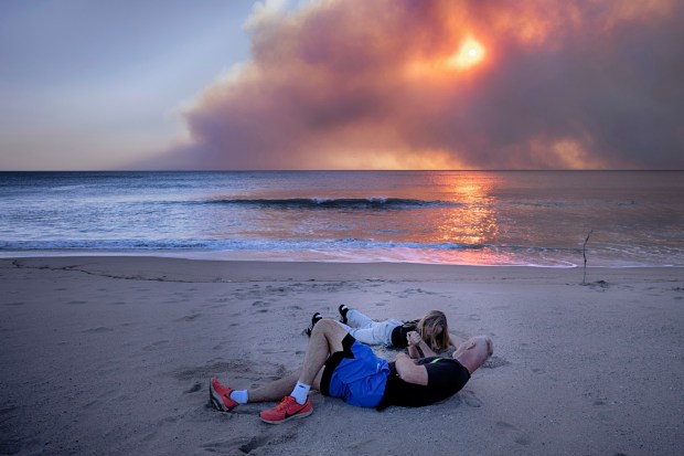 Tim Murphy and his son Sean arm wrestle on the beach during sunset under a blackened sky from a wildfire in the Pacific Palisades